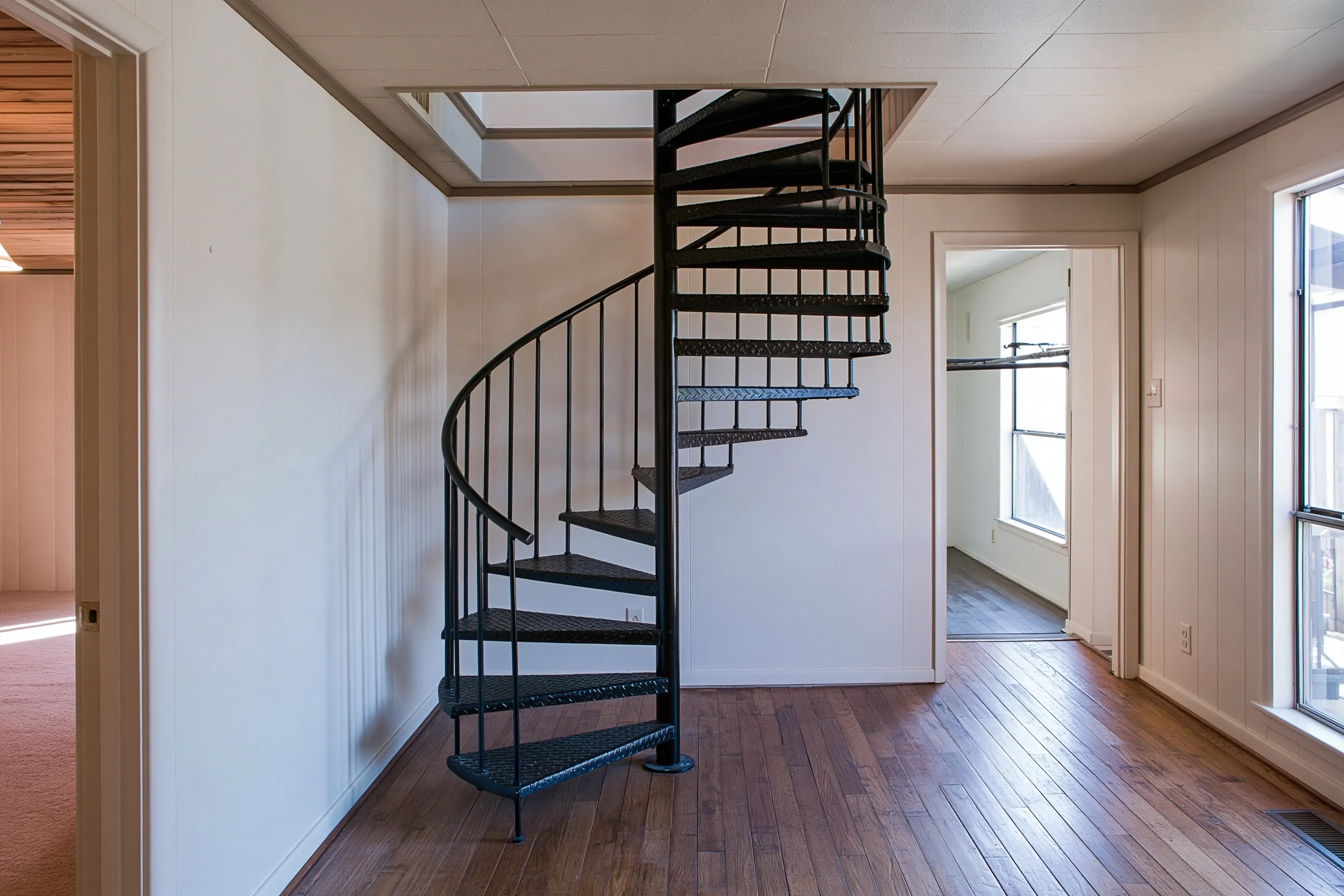 Compact black steel spiral stair with diamond-plate treads rising through a square opening in a paneled room
