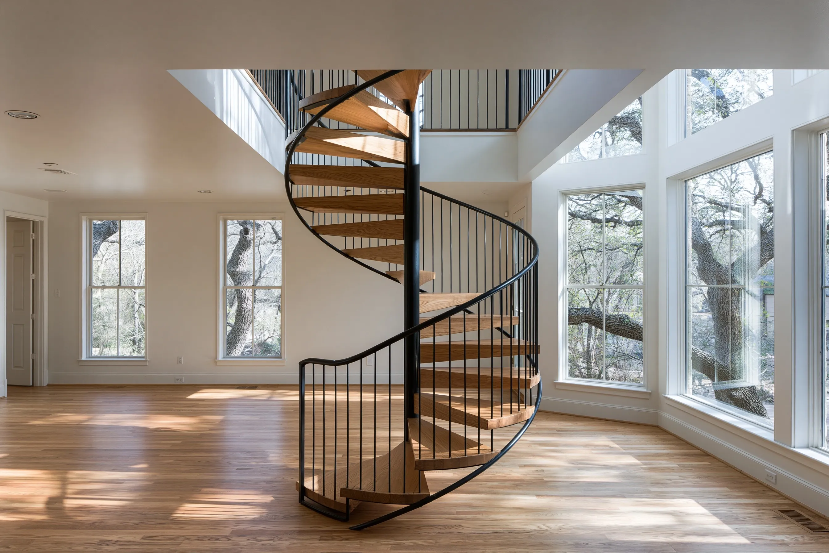Black steel spiral stair with oak treads rising to an upstairs walkway in a two-story room with tall windows